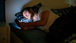 Un joven acostado en la cama por la noche y usando su teléfono inteligente mientras su novia duerme